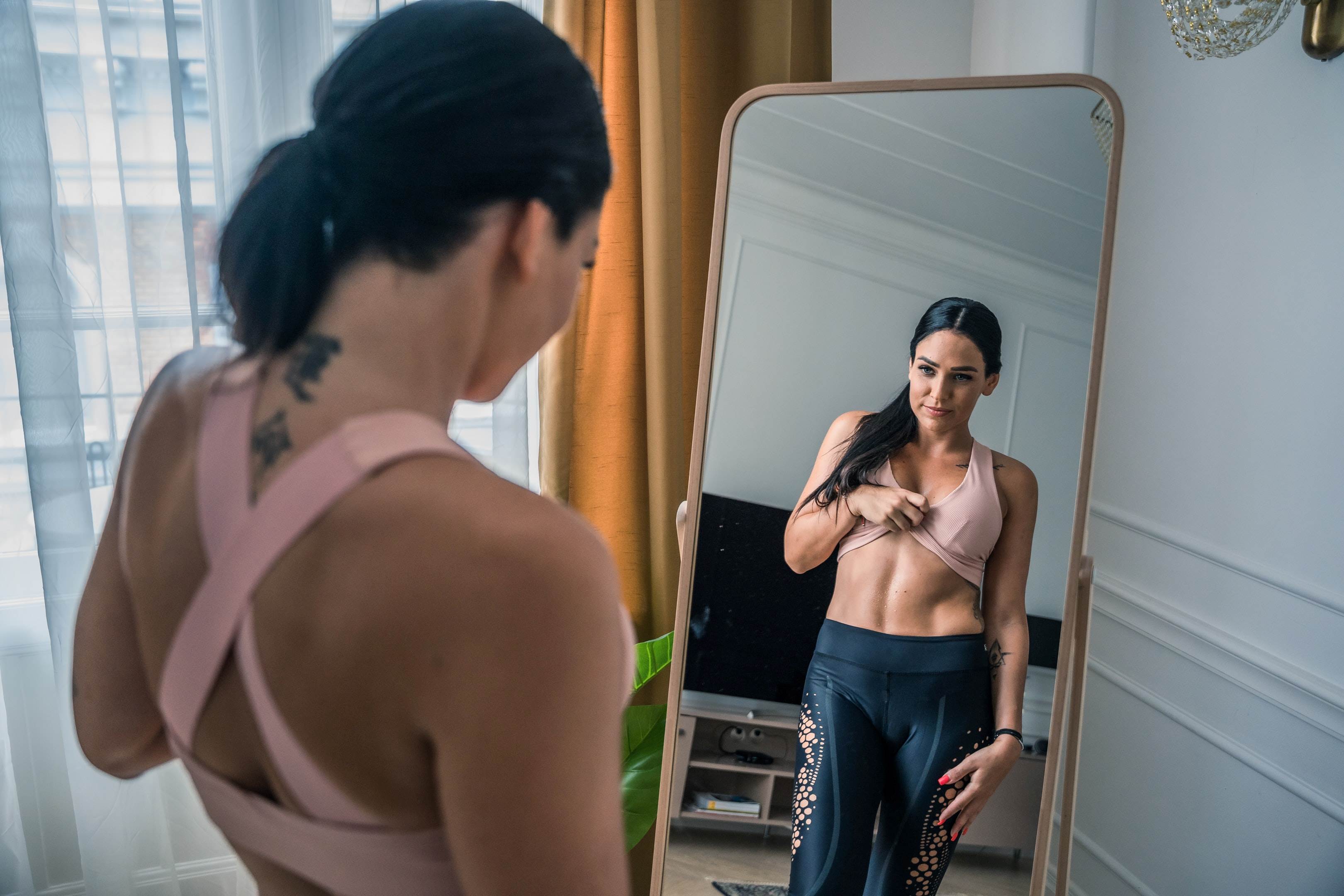 Žena ve fitness úboru stojí před zrcadlem. Částečně si vyhrnuje sportovní podprsenku a pozoruje svůj štíhlí pas.